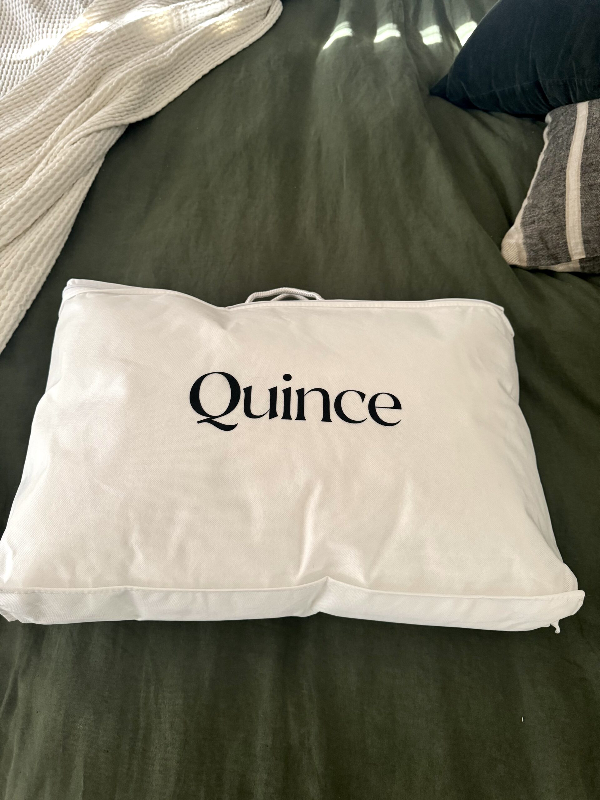 White bag with the brand name "Quince" printed on top, placed on a green bed with other linens partially visible.