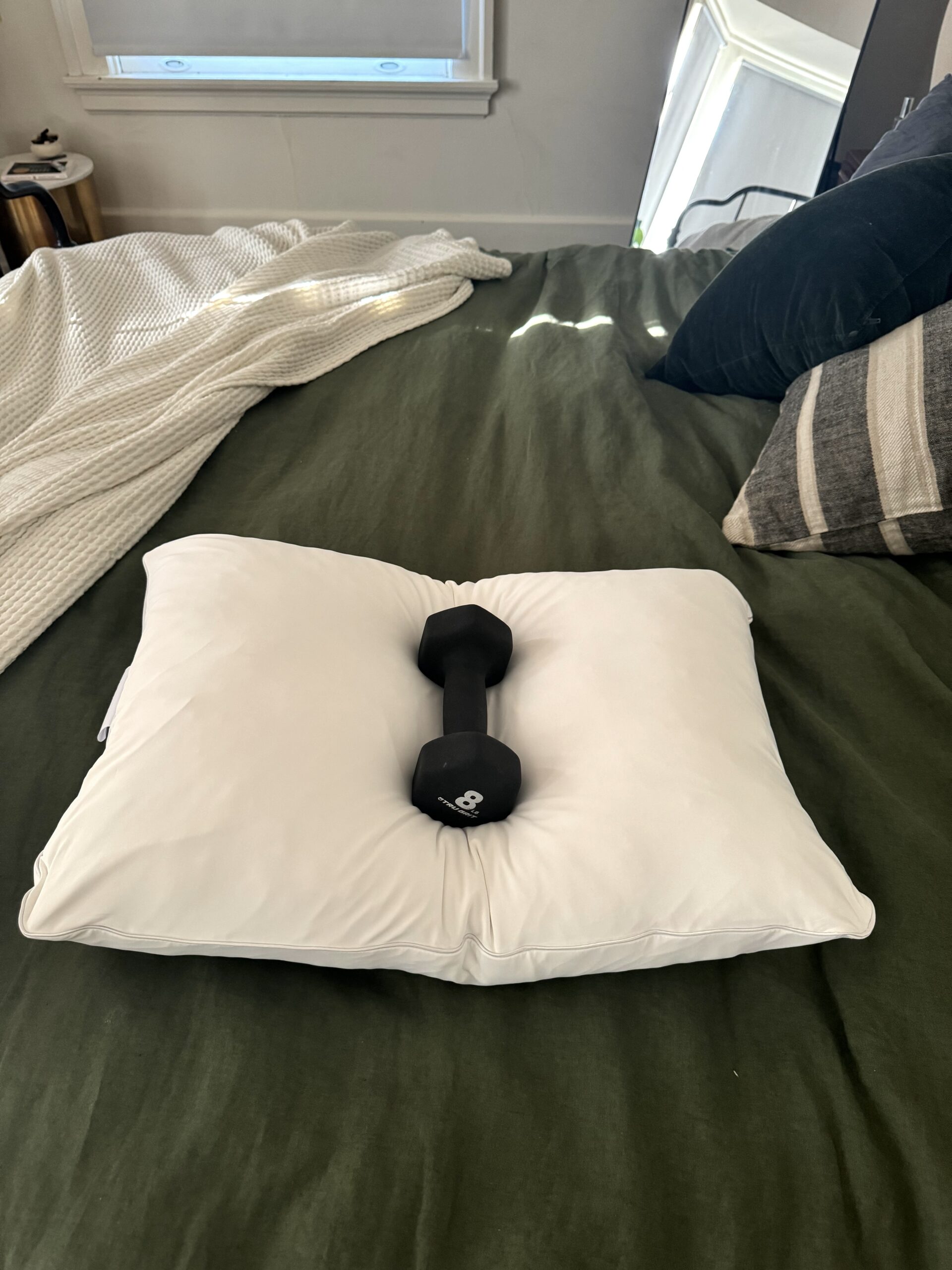 A single dumbbell rests on a white pillow placed on a green bedspread.