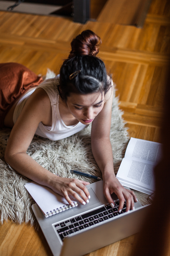 Person lying on a rug working on a laptop with an open notebook and book beside them on a wooden floor.