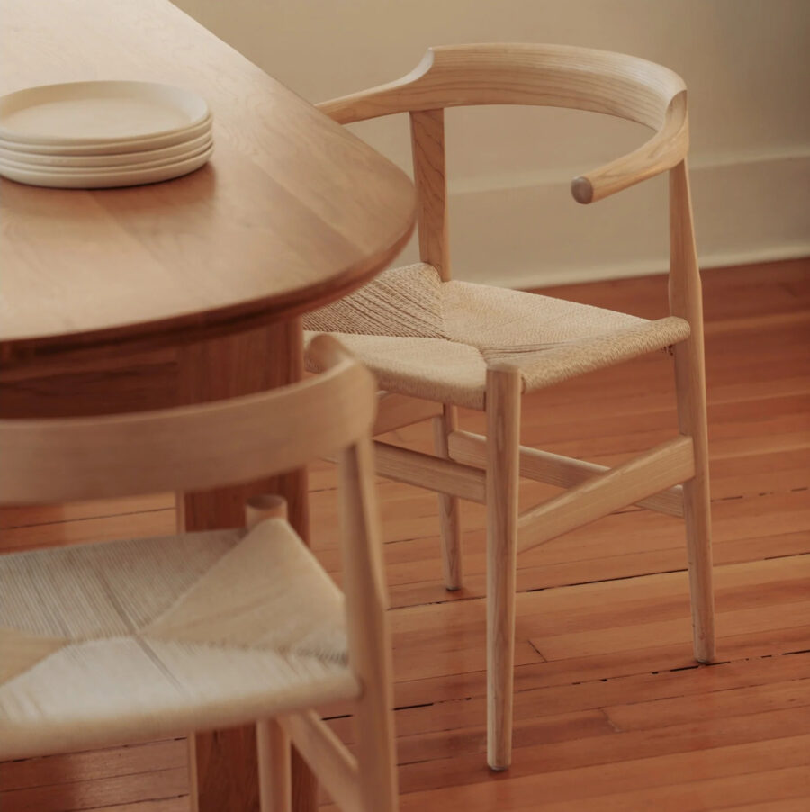 A wooden dining table with stacked white plates and a chair with a woven seat and curved backrest on a hardwood floor.