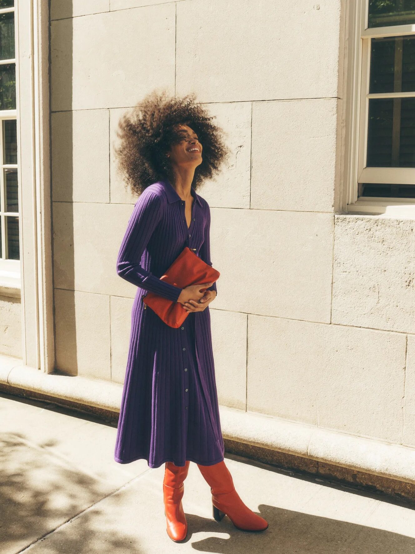 Person in a purple dress and red boots stands outside, holding a red clutch bag, looking upward with a smile.