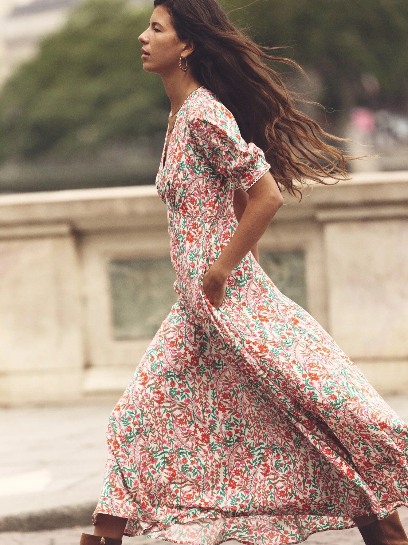 A woman with long, flowing hair walks outdoors, wearing a long floral dress with her hands in her pockets.