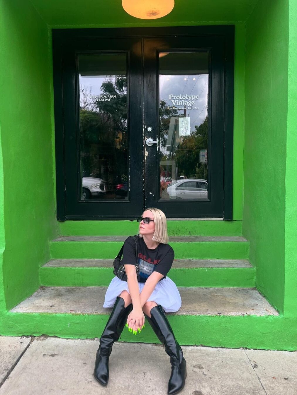 A person with blonde hair, wearing sunglasses, a black shirt, and black boots, sits on green steps in front of a black door labeled "Prototype Vintage.