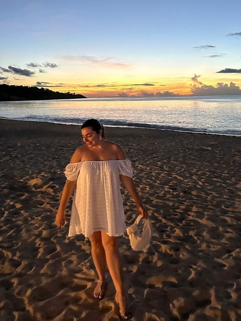 A person in a white off-shoulder dress walks on a sandy beach at sunset, with the ocean and clouds in the background.