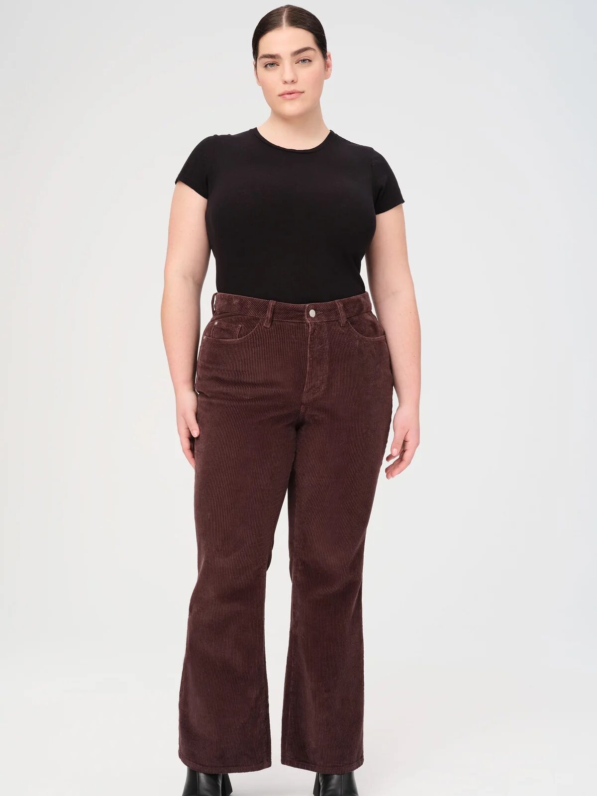 A person stands against a plain background, wearing a black t-shirt, brown corduroy pants, and black ankle boots.