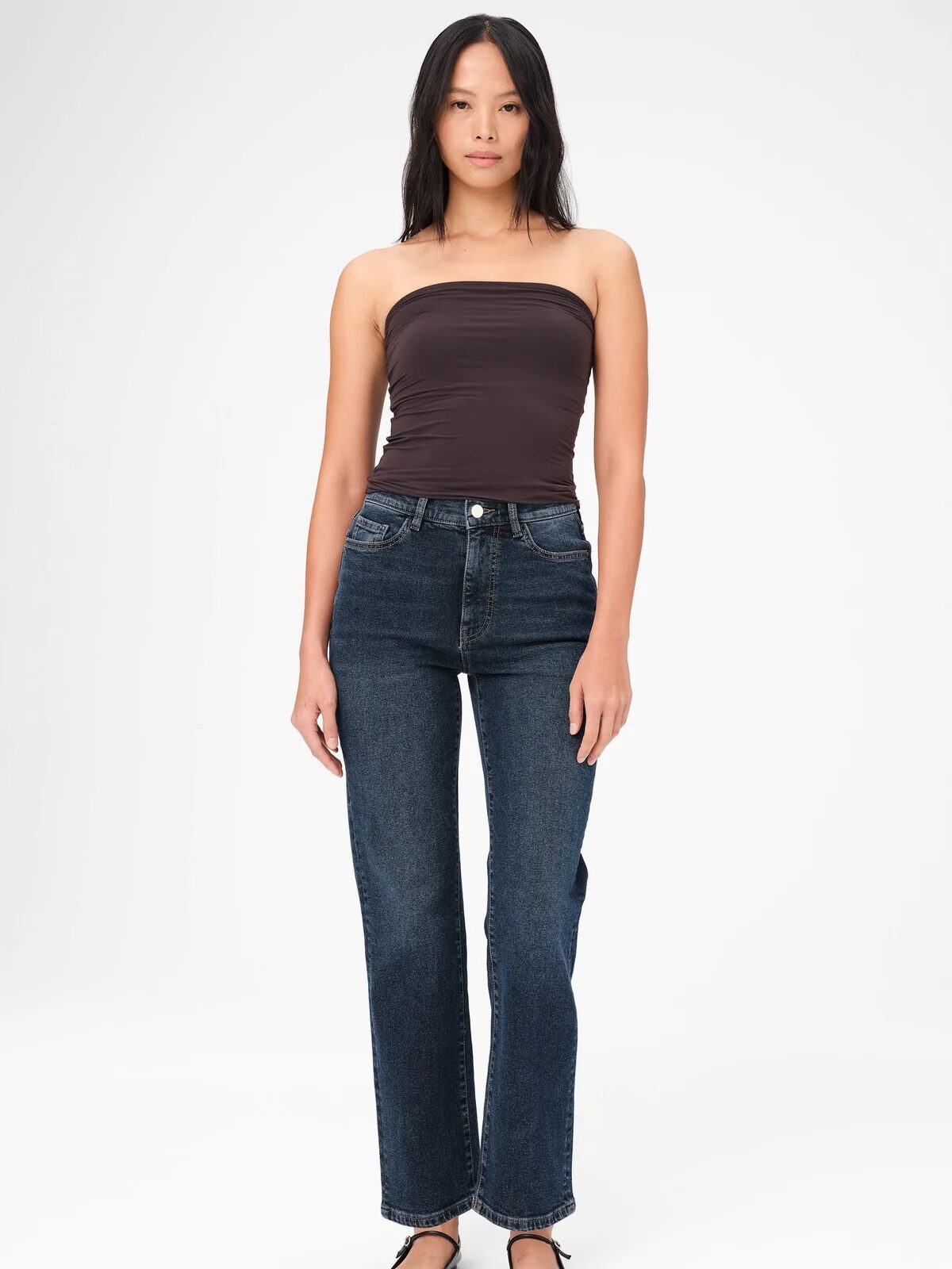 A woman stands against a plain background, wearing a dark strapless top, blue jeans, and black ballet flats.