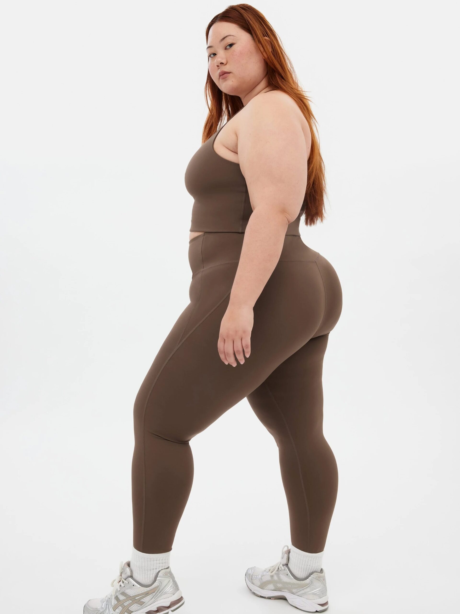 A plus size model wearing a matching brown athleisure tank and leggings from Girlfriend Collective. 