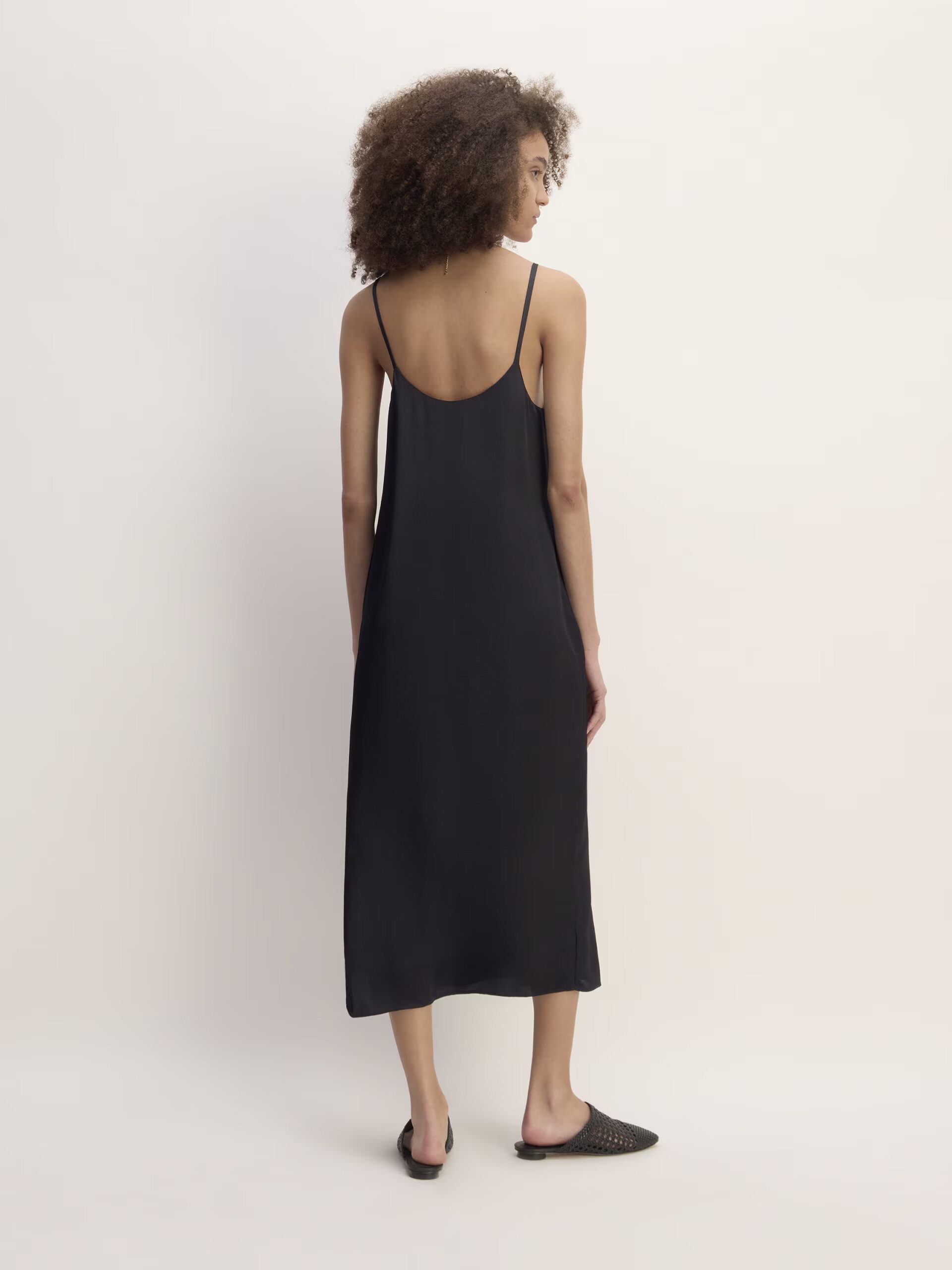 A person with curly hair is standing with their back facing the camera, wearing a black sleeveless dress and black slip-on shoes against a plain background.