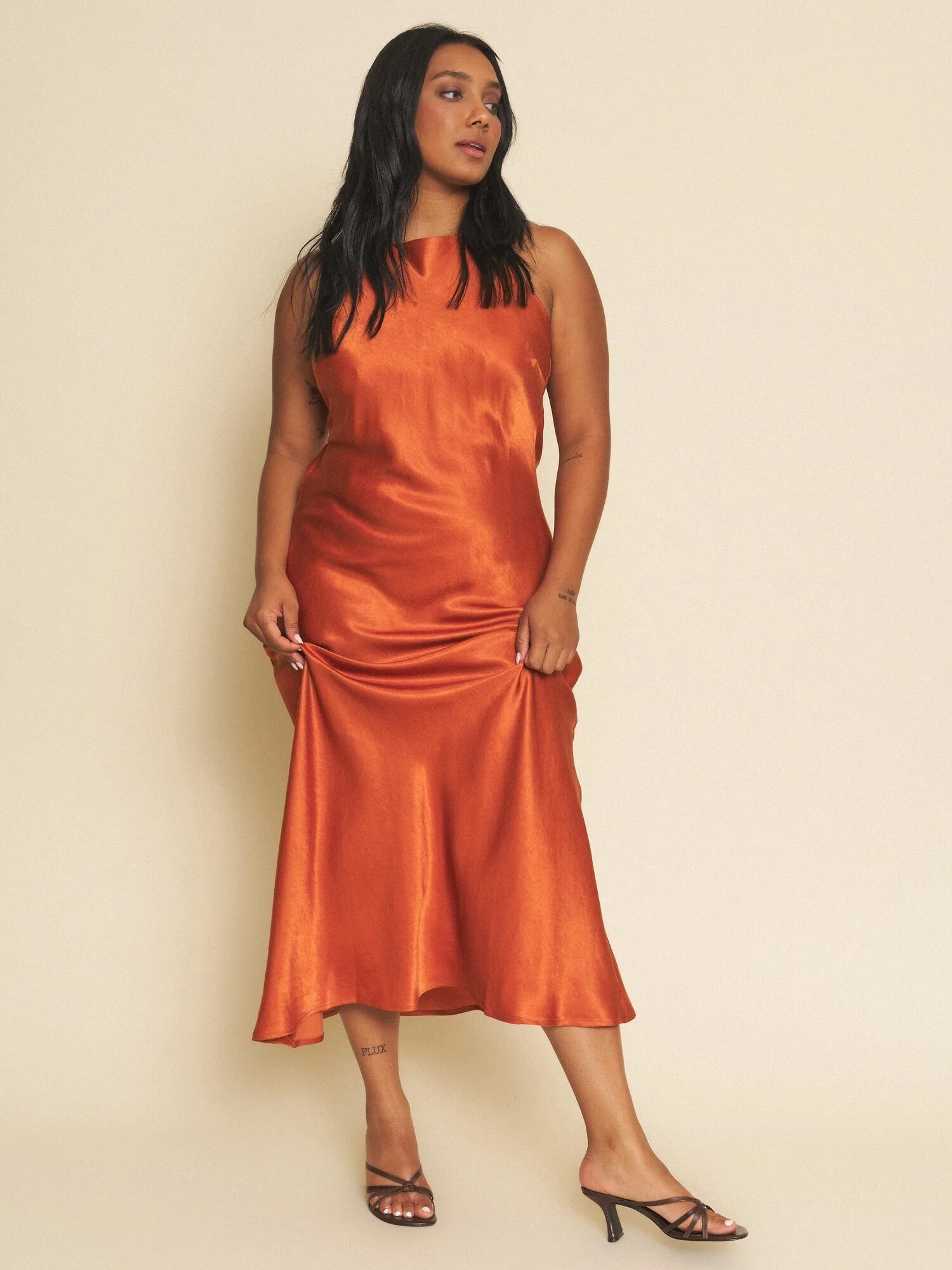 A woman stands against a plain background wearing an orange satin sleeveless dress and black heeled sandals.