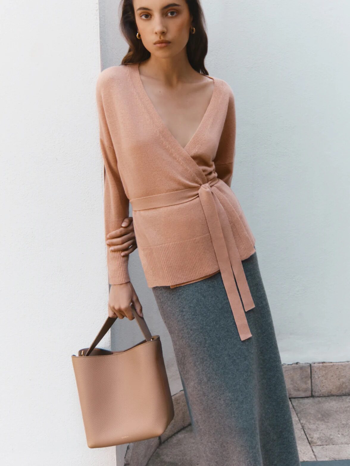 A woman wearing a beige wrap sweater and grey skirt holds a beige tote bag while standing against a white wall.