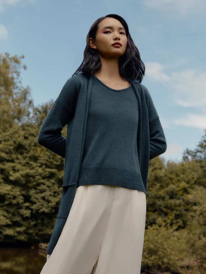 A woman with dark hair stands outdoors wearing a blue-gray sweater and cream pants, with trees and a cloudy sky in the background.