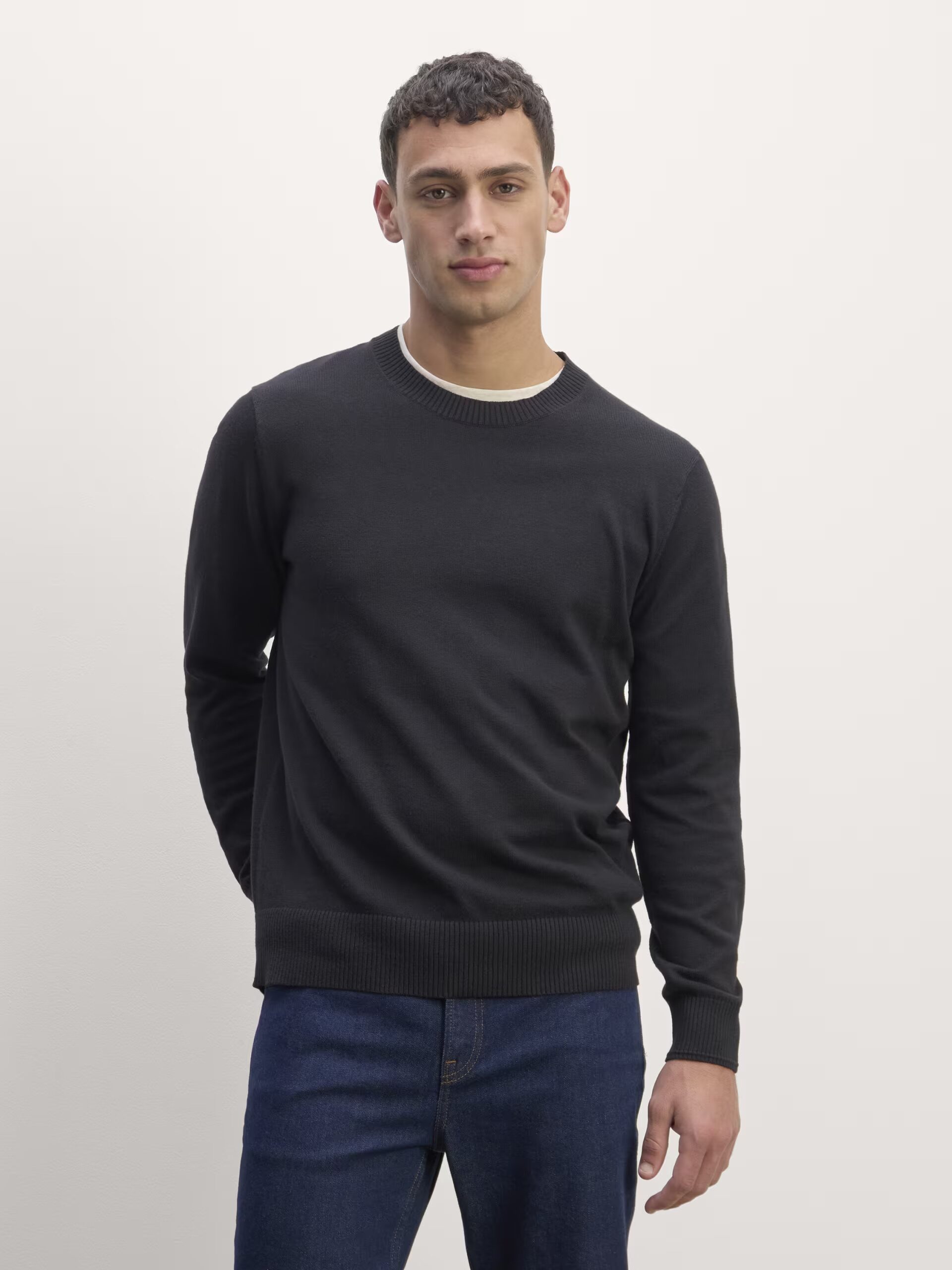 A man stands against a plain background, wearing a black sweater and blue jeans. He has short hair and a neutral expression.