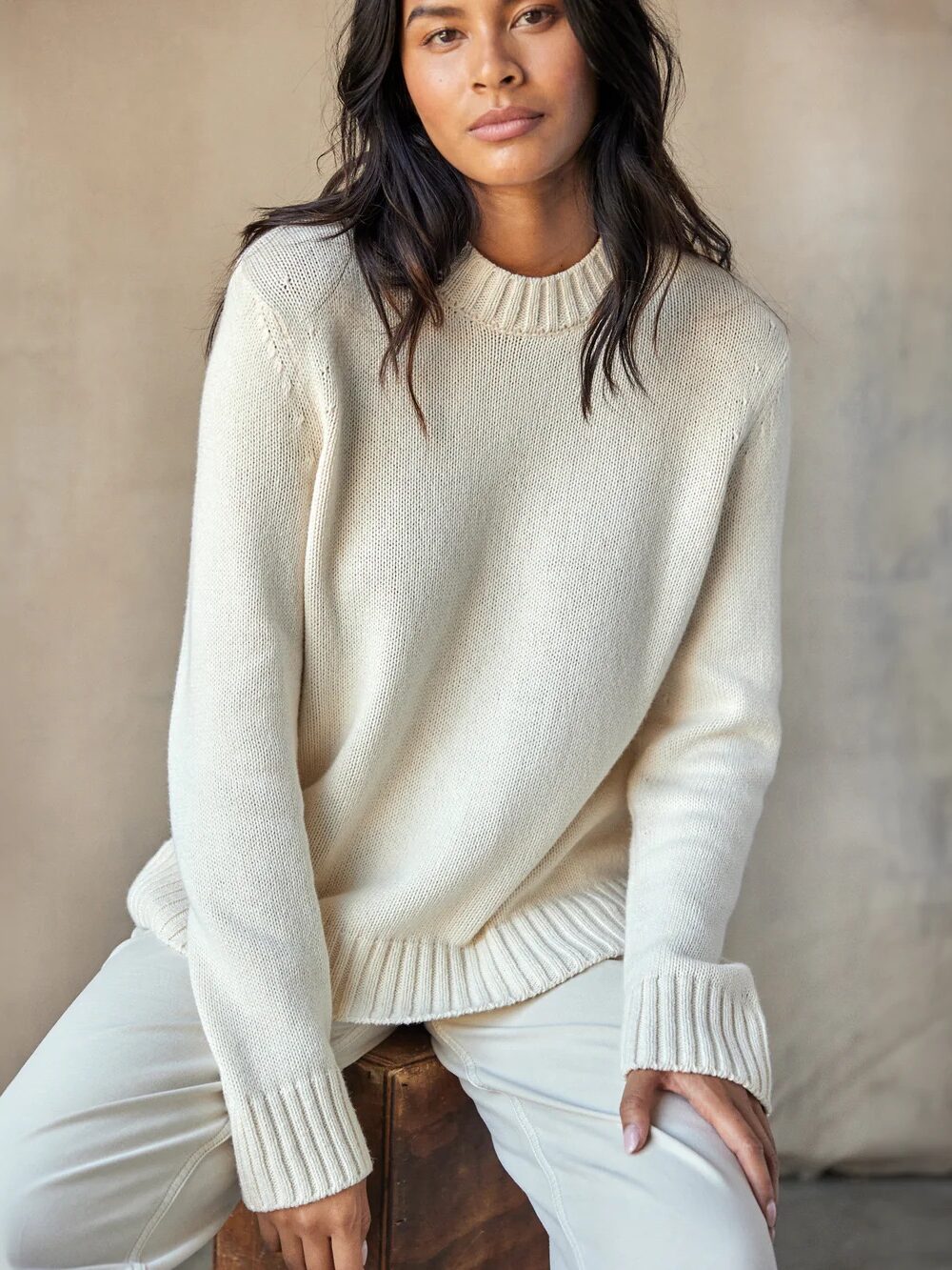 A person with long hair is wearing a light-colored sweater and pants while looking at the camera.