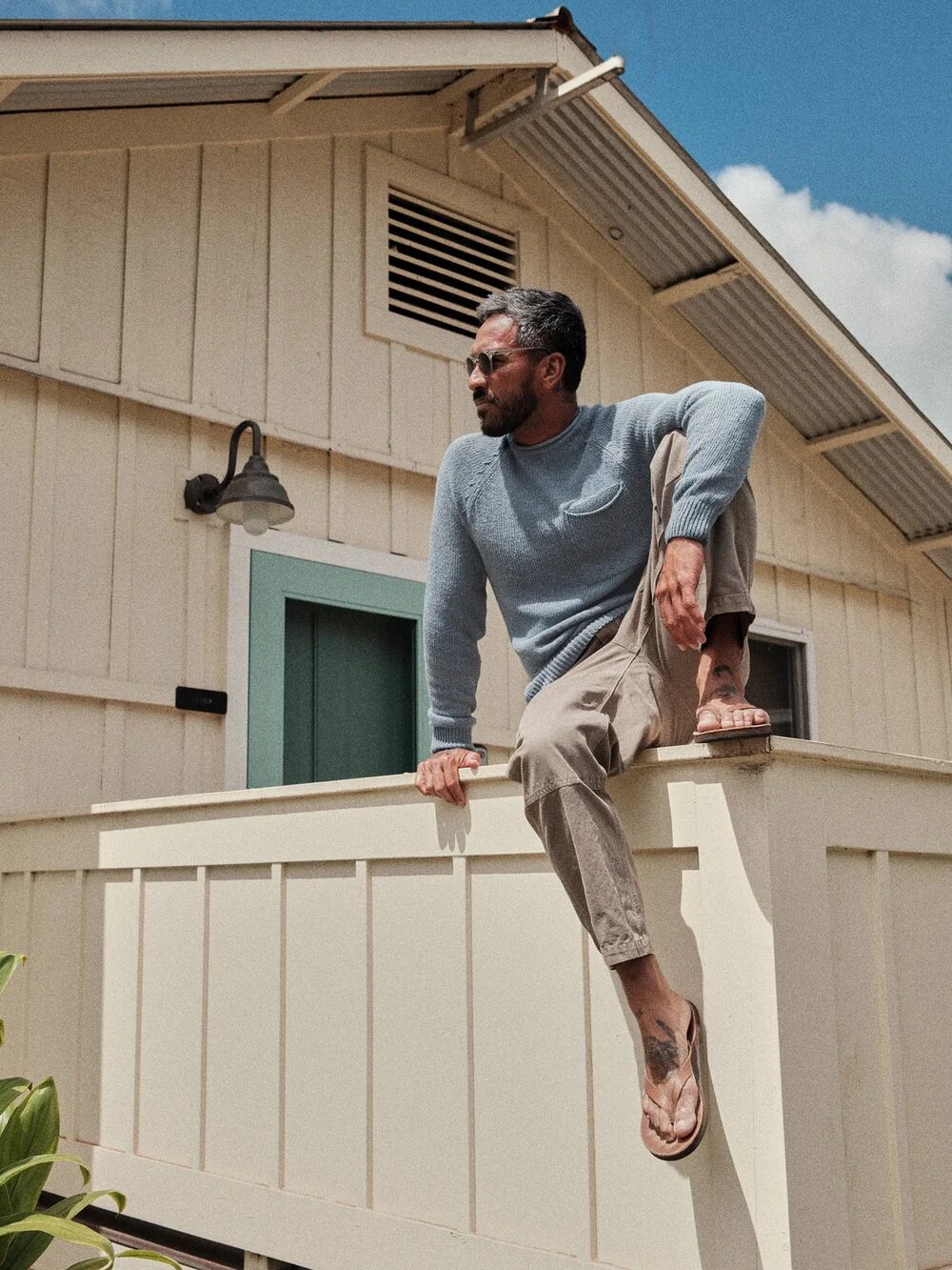 A man in casual attire sits on the railing of a beige wooden building, wearing sunglasses and sandals, looking to his left.