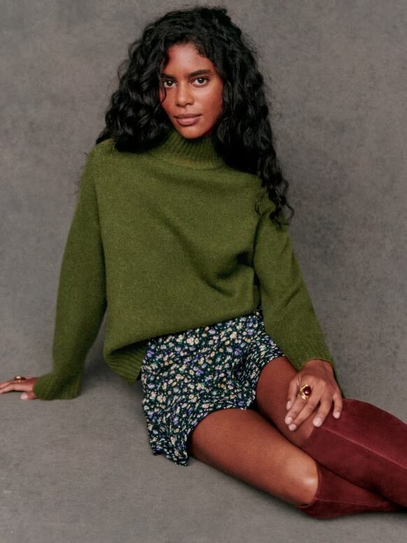 A person with long curly hair is sitting on the floor wearing a green sweater, a floral skirt, and maroon knee-high boots.