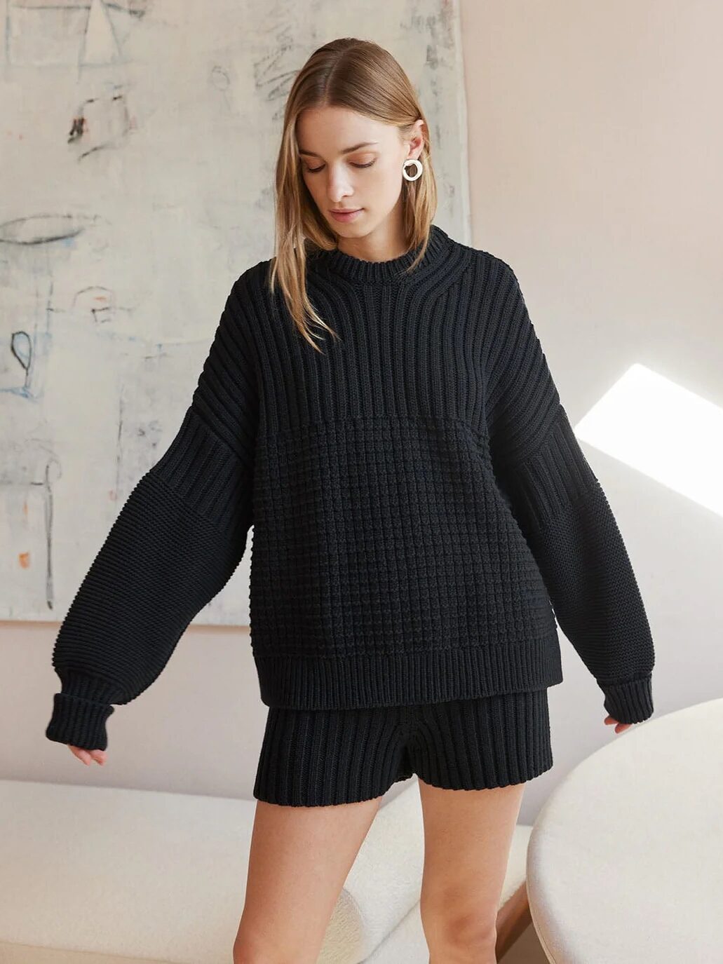 A woman with long hair wears a black, ribbed knit sweater and black knit shorts. She stands indoors in front of a textured wall with light abstract art.