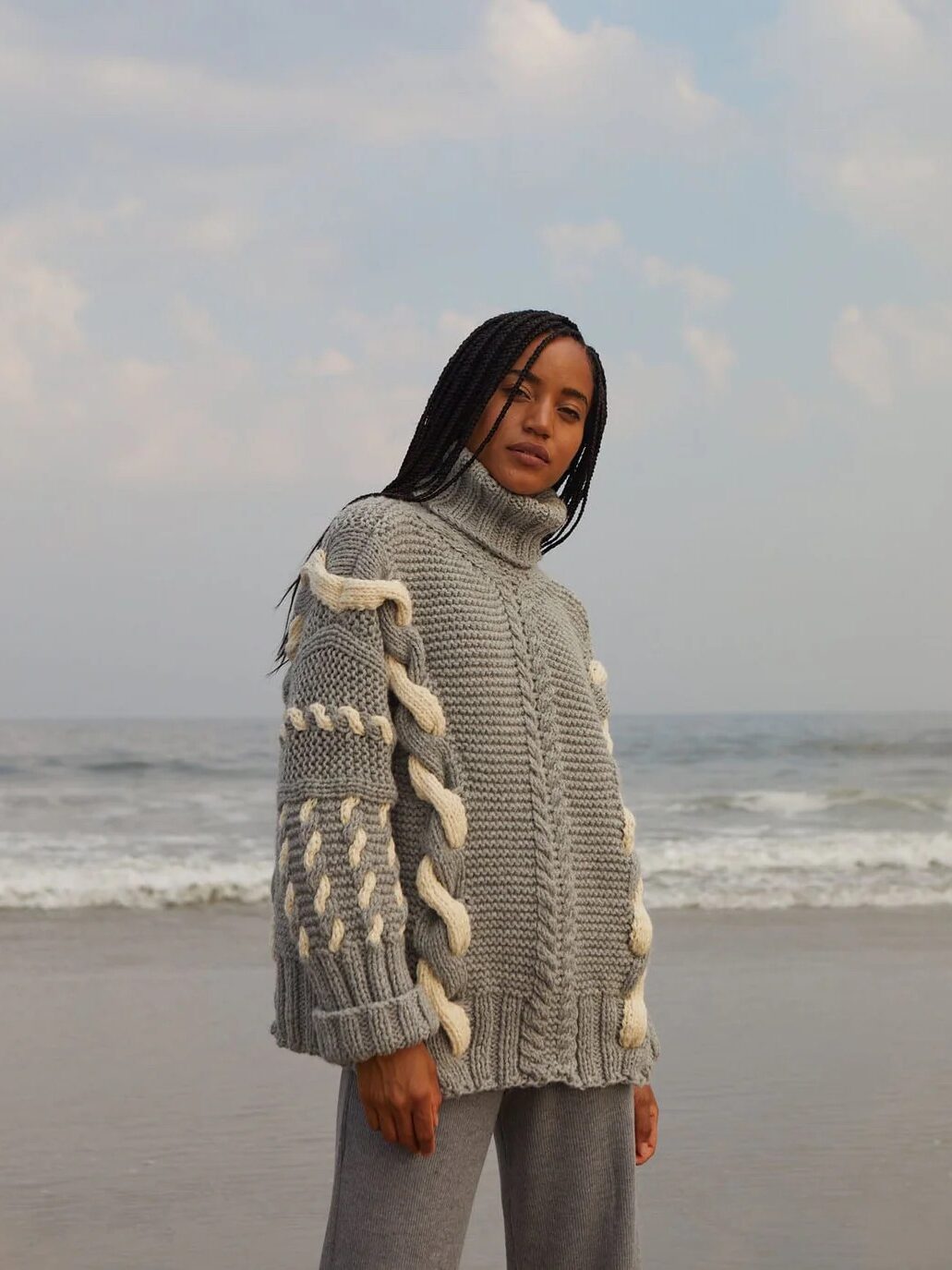 A person wearing a thick, gray cable knit sweater and gray pants stands on a beach with the ocean and cloudy sky in the background.