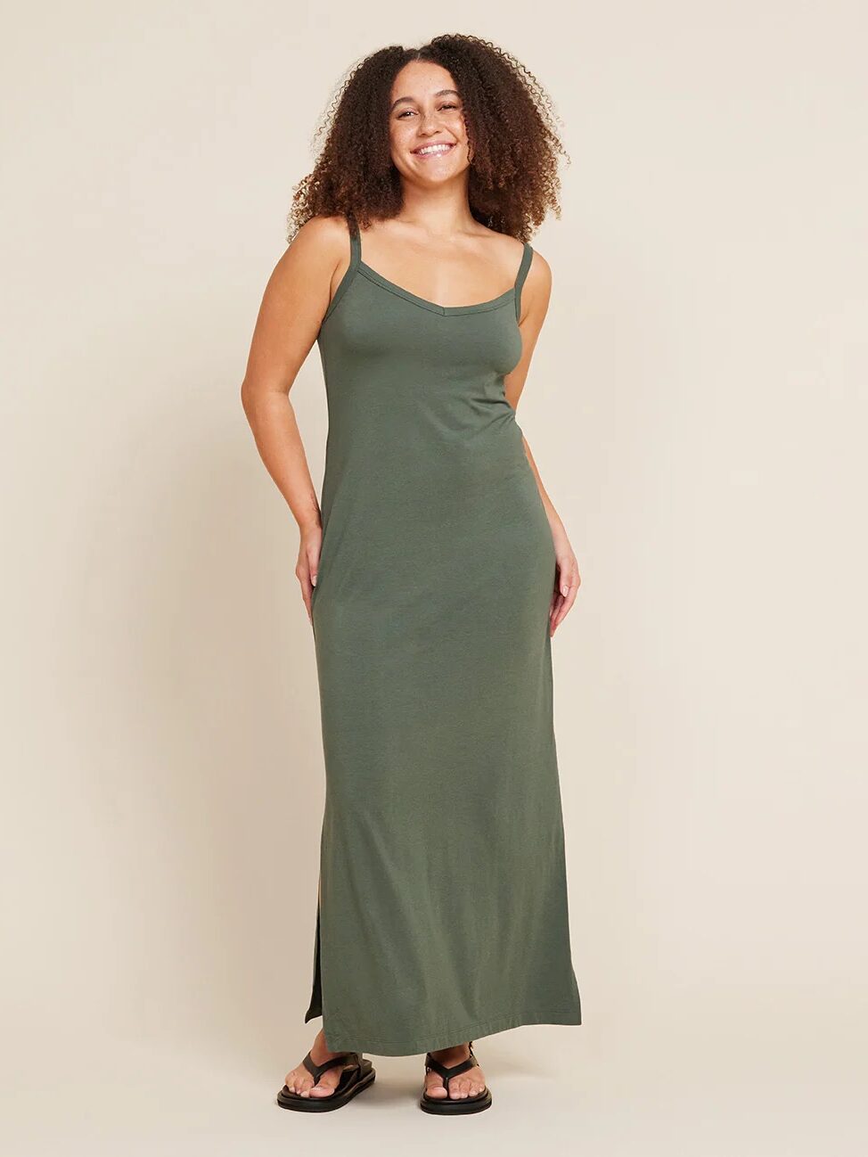 A woman in a green maxi dress with spaghetti straps stands against a neutral background, smiling with her hands at her sides. She is wearing black sandals.