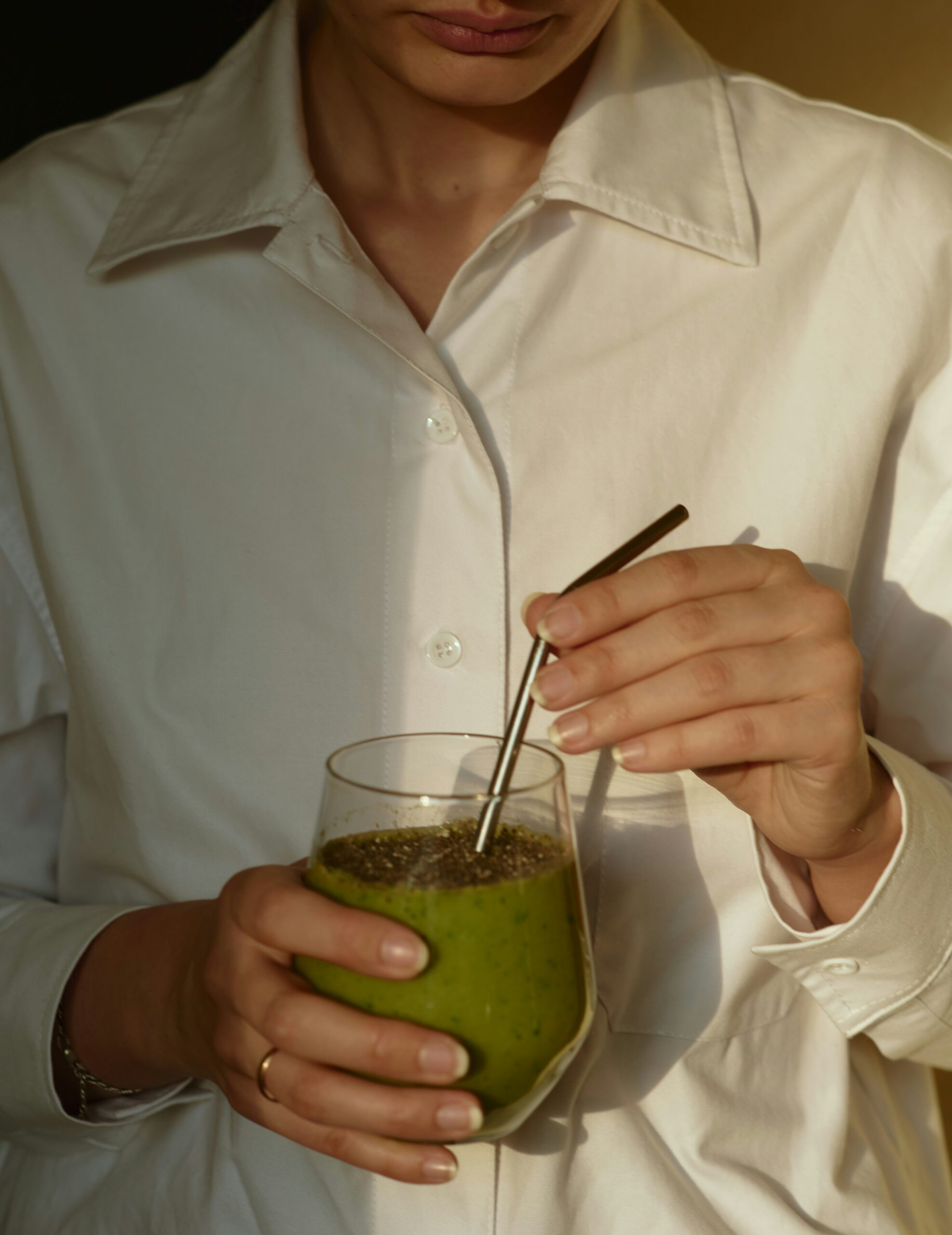 A person in a white shirt stirs a green smoothie in a clear glass with a metal straw.