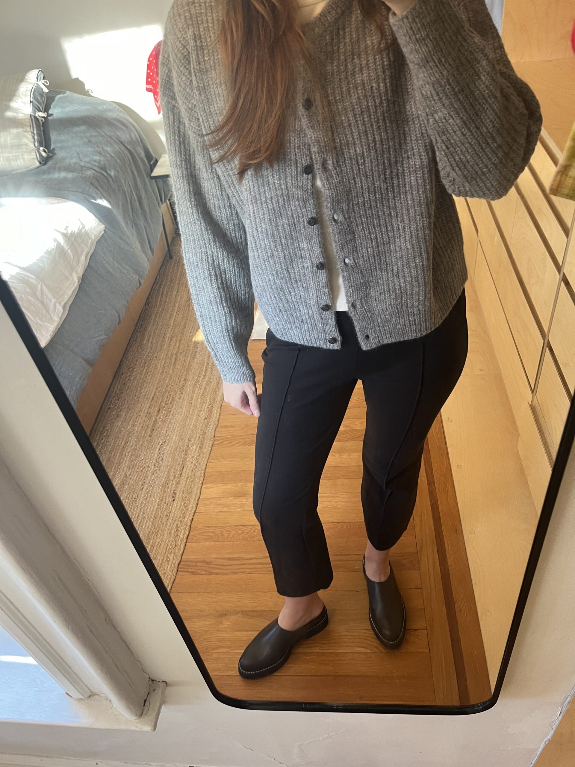 A woman in black pants, black shoes, and a gray cardigan taking a photo of her clothes in a mirror.