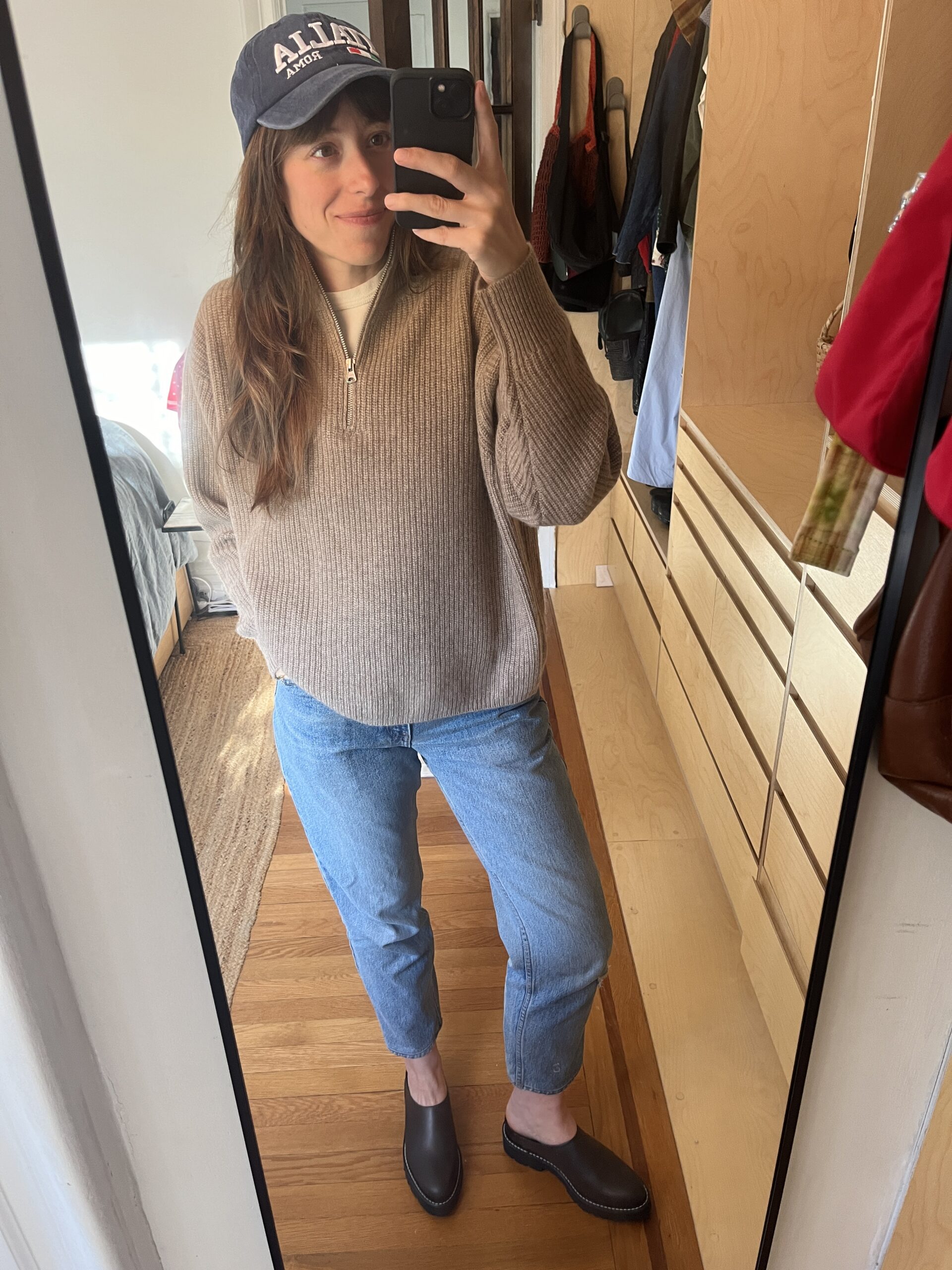 A woman in a baseball hat, oatmeal colored half-zip sweater, jeans, and dark shoes takes a photo of herself in a full-length mirror.