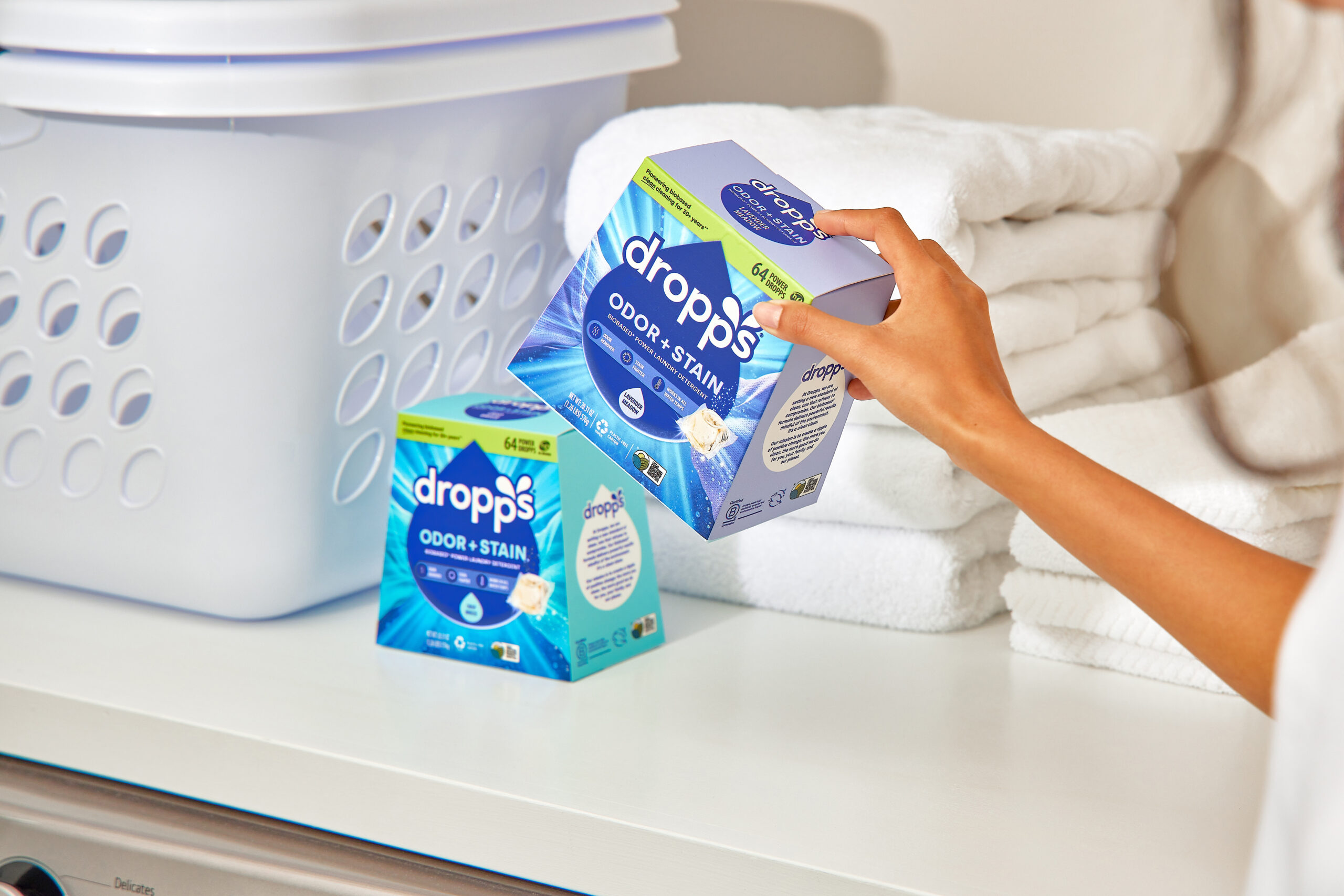 Two boxes of Dropps Odor and Stain laundry pods set nearby a stack of clean white towels and a laundry basket. 