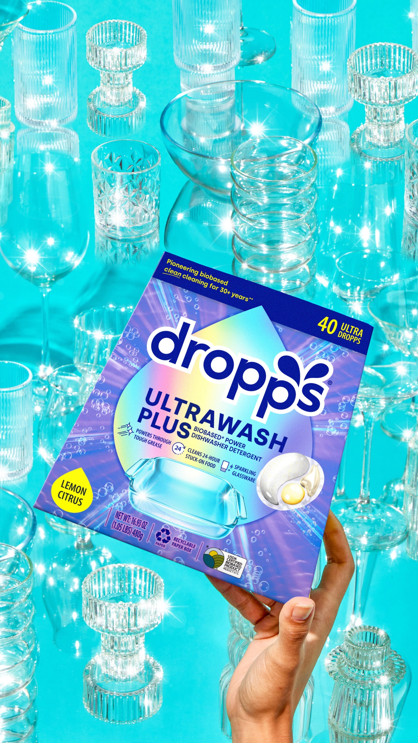 A box of 40 Dropps Ultrawash Plus Dishwasher pods held up in front of many clean glasses on a blue background