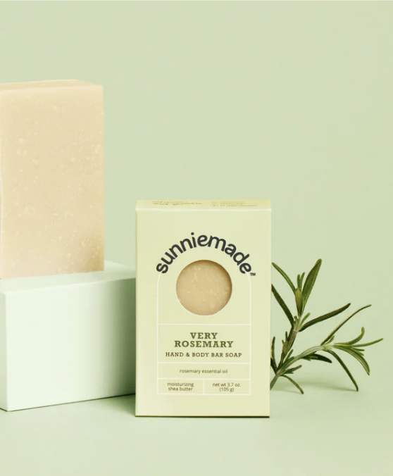 A box and a bar of Sunnie Made Very Rosemary hand and body soap are placed on a light green surface next to a sprig of rosemary.