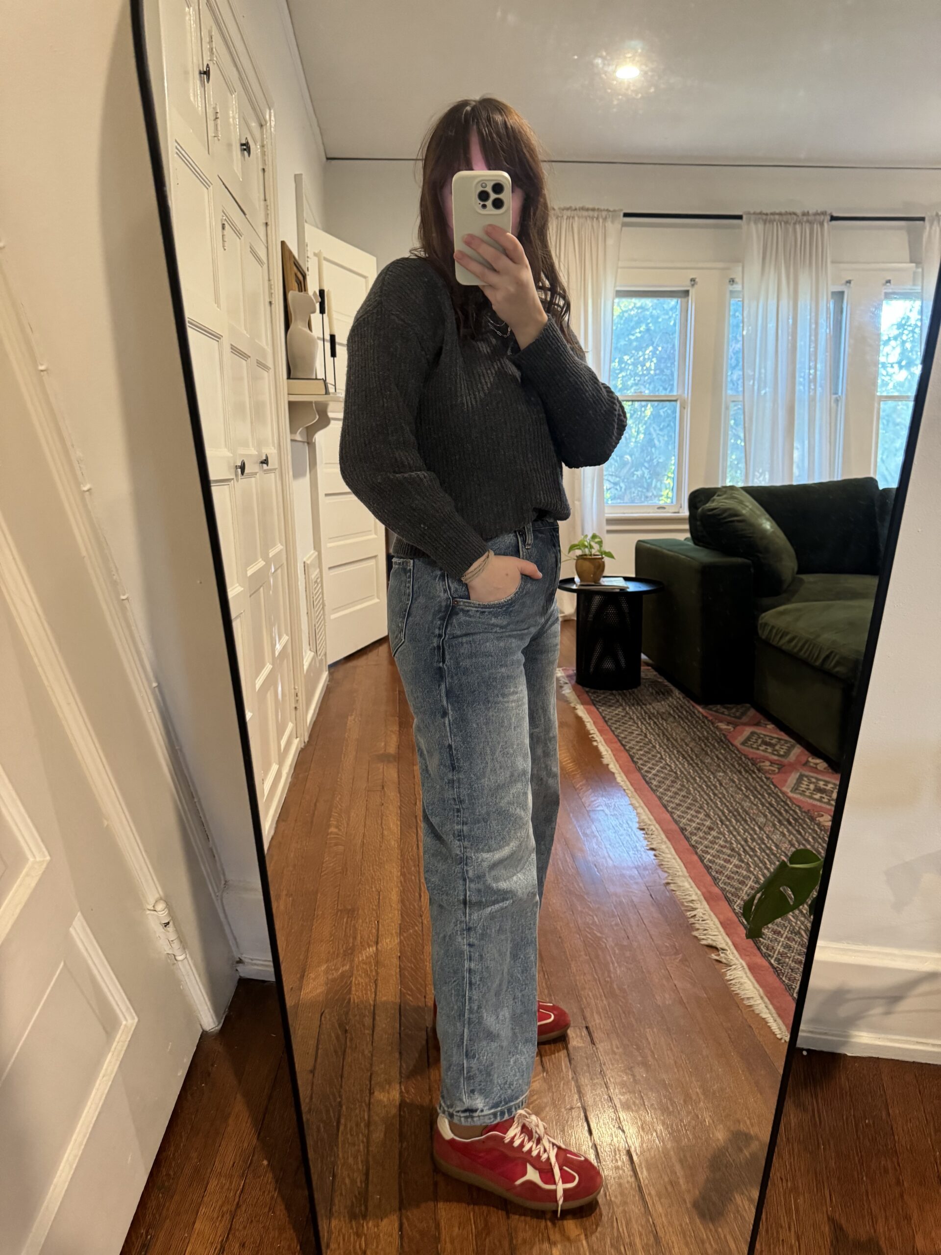 Person taking a mirror selfie wearing a dark sweater, light blue jeans, and red sneakers in a living room with wooden floor and white walls.