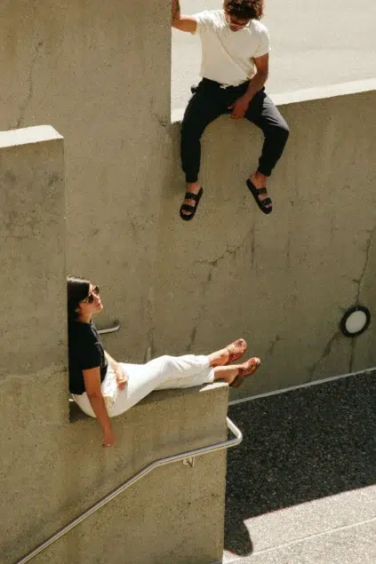 Two people sit on separate levels of a concrete structure, with one person sitting higher. They are casually dressed and are outdoors in sunlight.