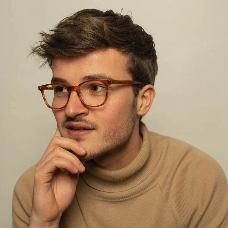 A person with short hair and glasses wearing a beige turtleneck gazes thoughtfully to the side.