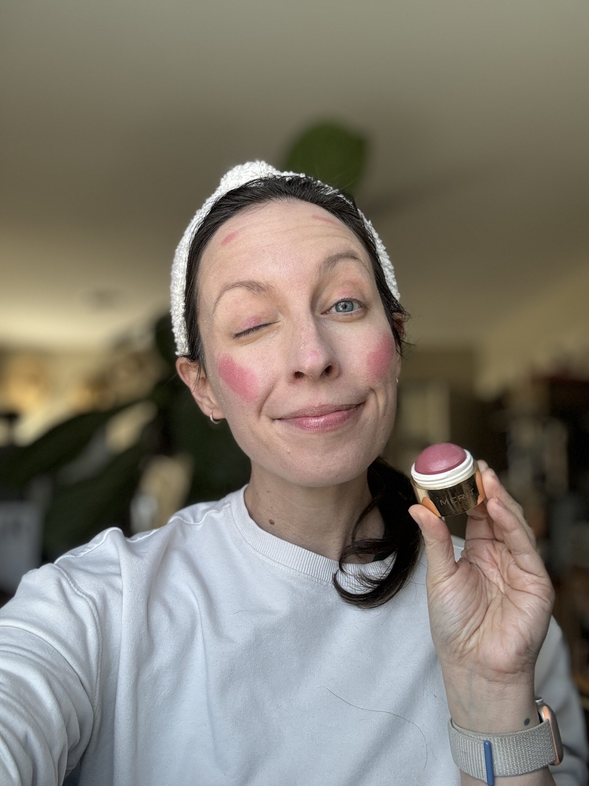 Person wearing a white headband and shirt applies blush to their cheeks, smiling with one eye closed, holding a small container of pink blush.