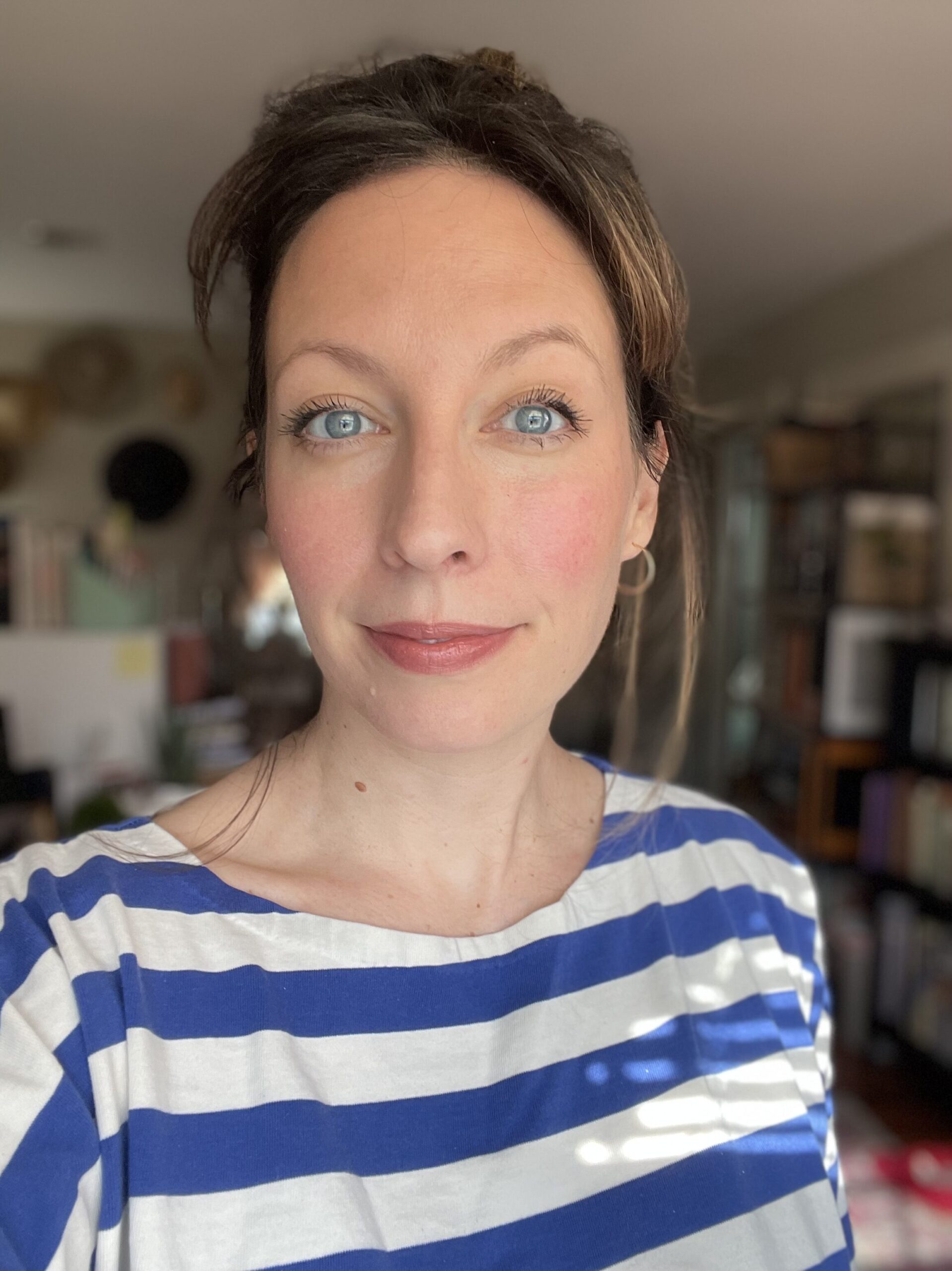 A woman with blue eyes, wearing a blue and white striped shirt, stands indoors.