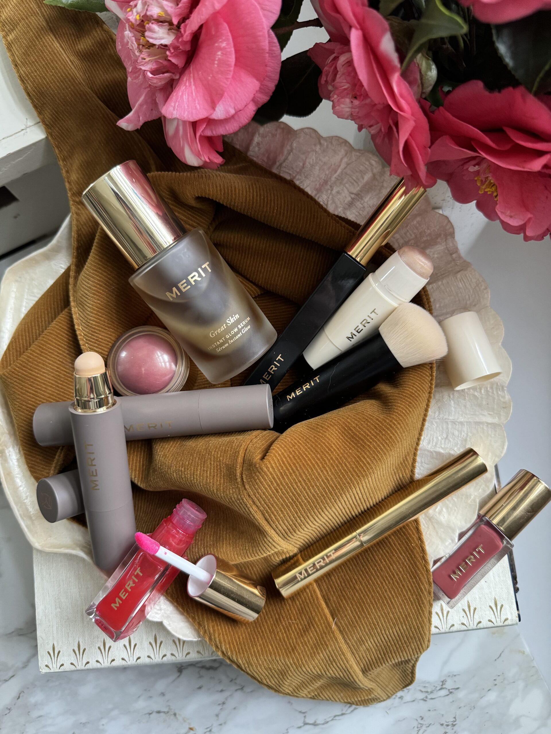 A collection of Merit beauty products, including foundation, blush, mascara, and lipstick, arranged on a brown fabric with pink flowers in the background.