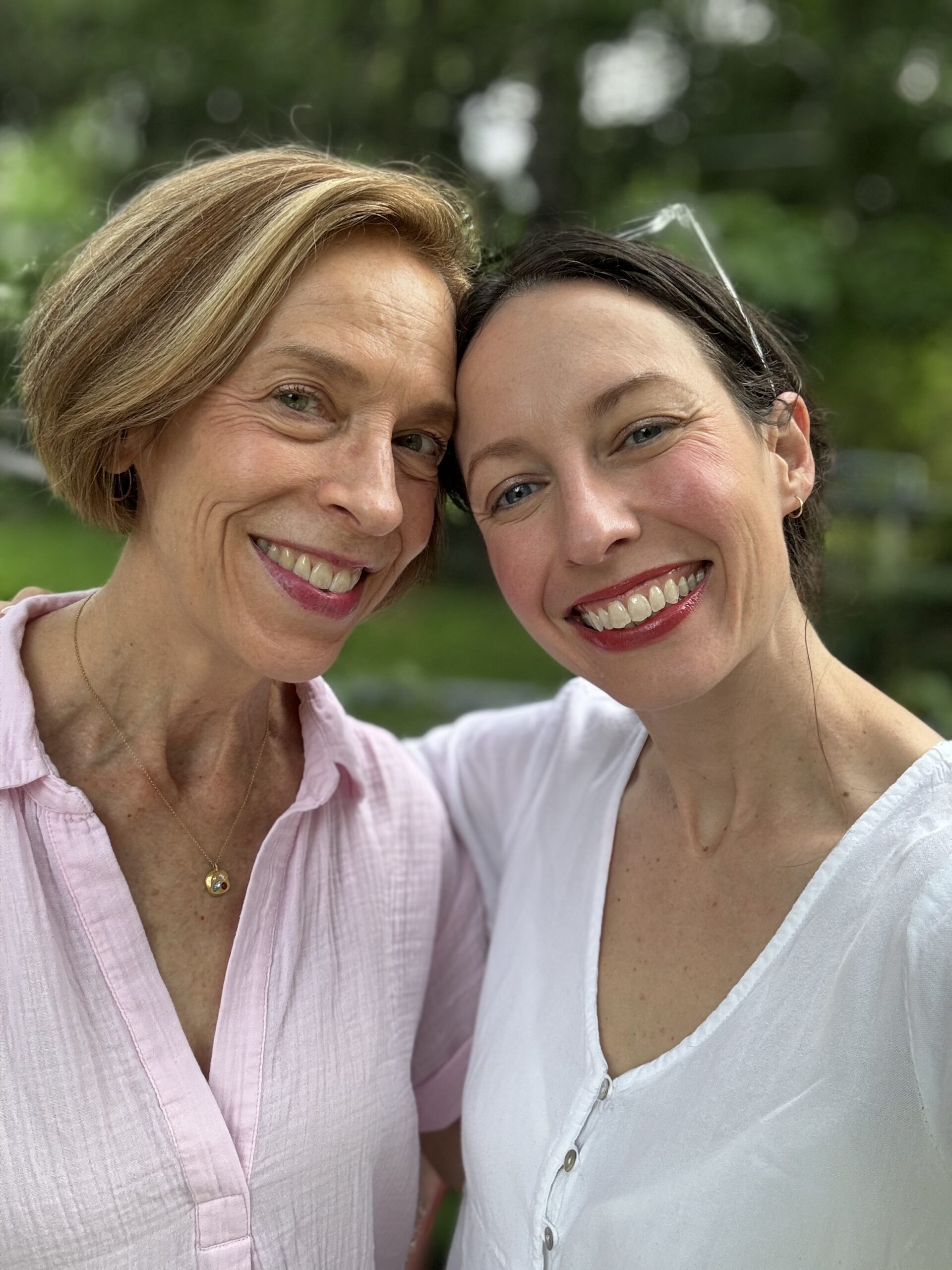 Two people smiling outdoors, standing close together, wearing light-colored shirts, with greenery in the background.