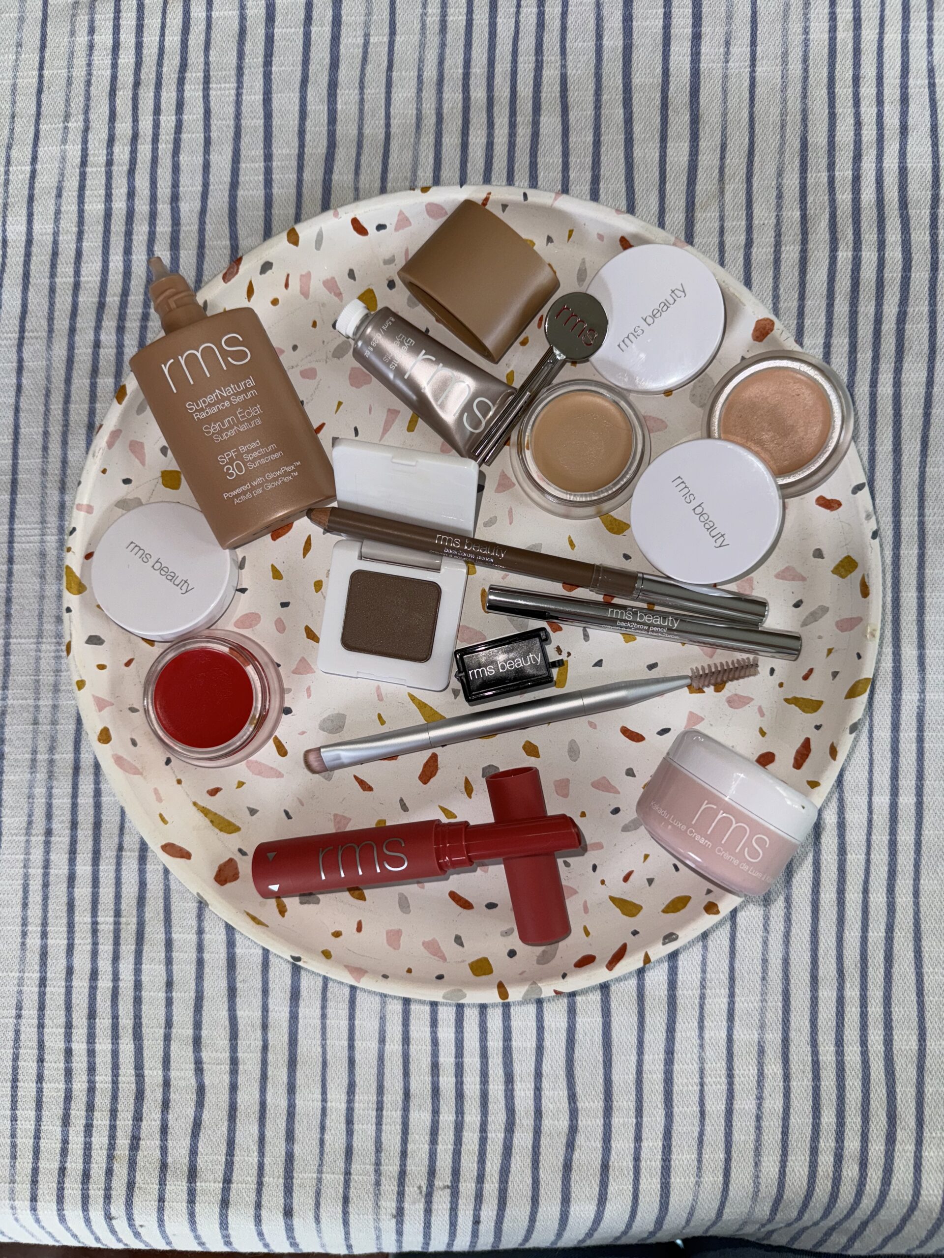 A variety of RMS beauty products arranged on a speckled tray with a striped cloth background, including lip, eye, and face cosmetics.