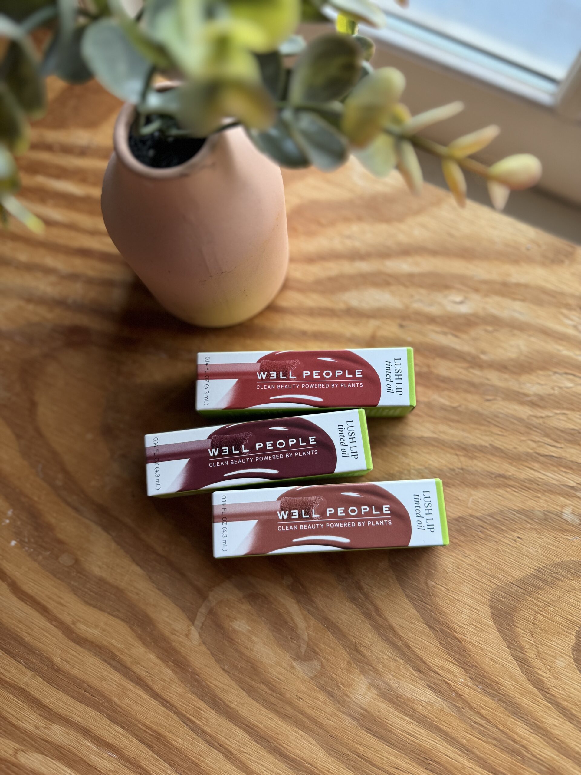 Three Well People beauty product boxes placed on a wooden surface near a pink vase with artificial greenery.