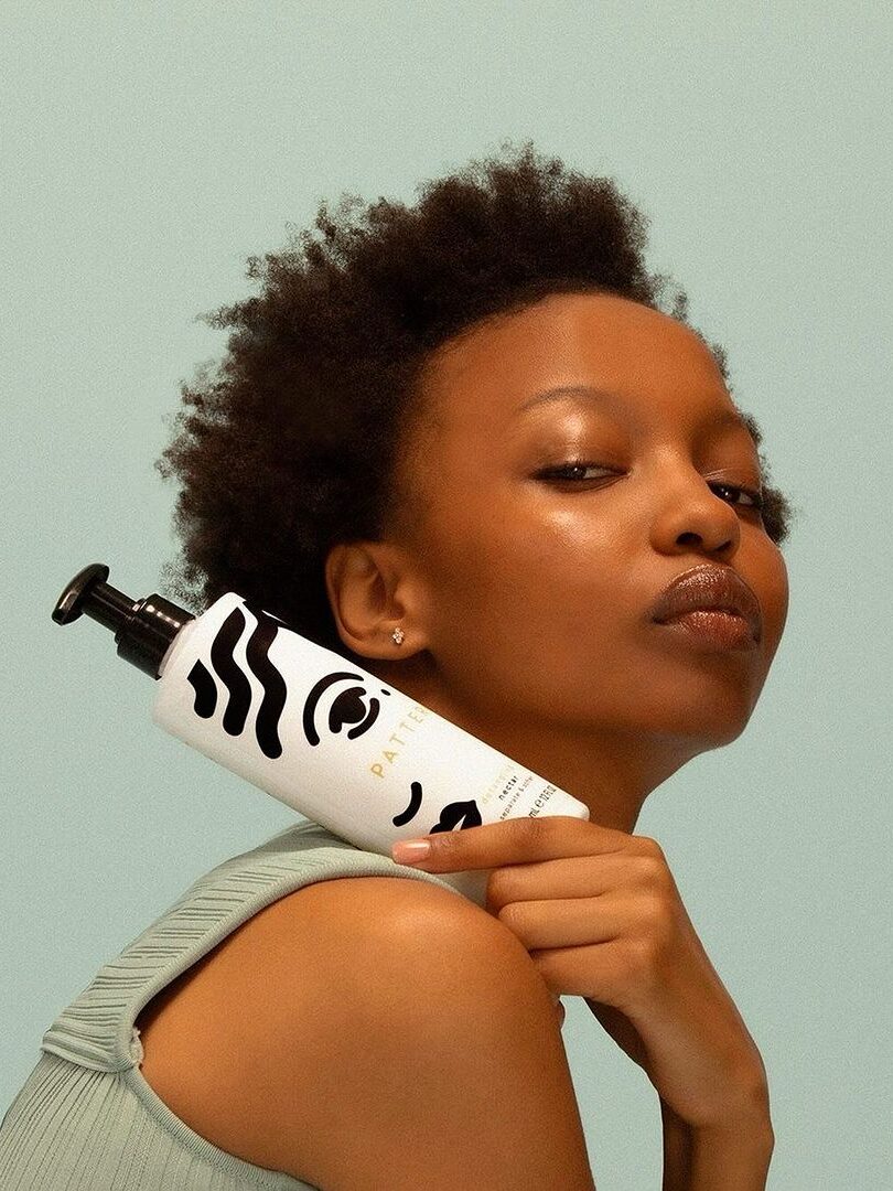 A curly-hair model holding up a bottle of PATTERN Beauty's detangling nectar over their shoulder. 