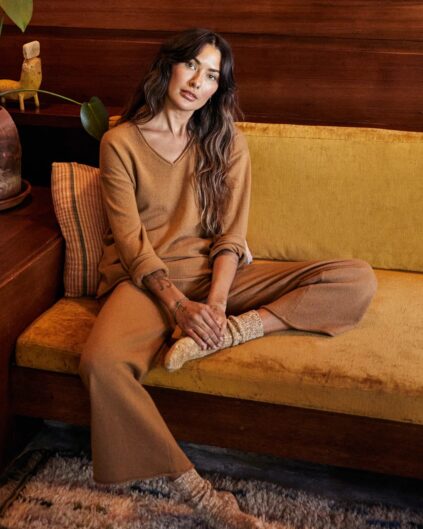 A person with long hair sits casually on a yellow couch, wearing a brown outfit and knitted socks, in a warmly lit room.