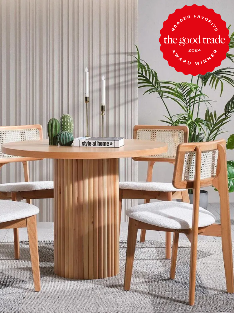 Modern dining area with a round wooden table, four chairs with rattan backs, and two candles in holders. A potted plant is in the background. "The Good Trade" award logo is in the corner.