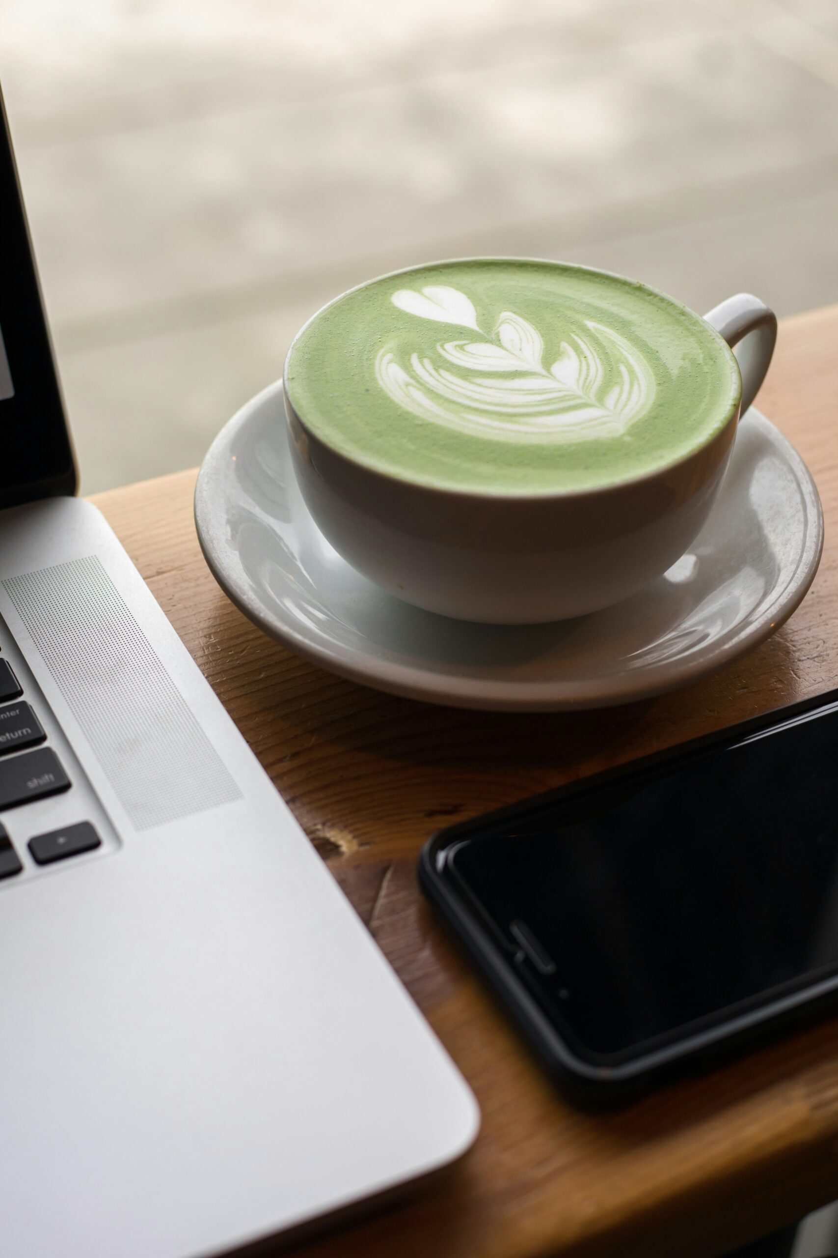 A cup of matcha latte with foam art sits on a wooden table next to a smartphone and a laptop.