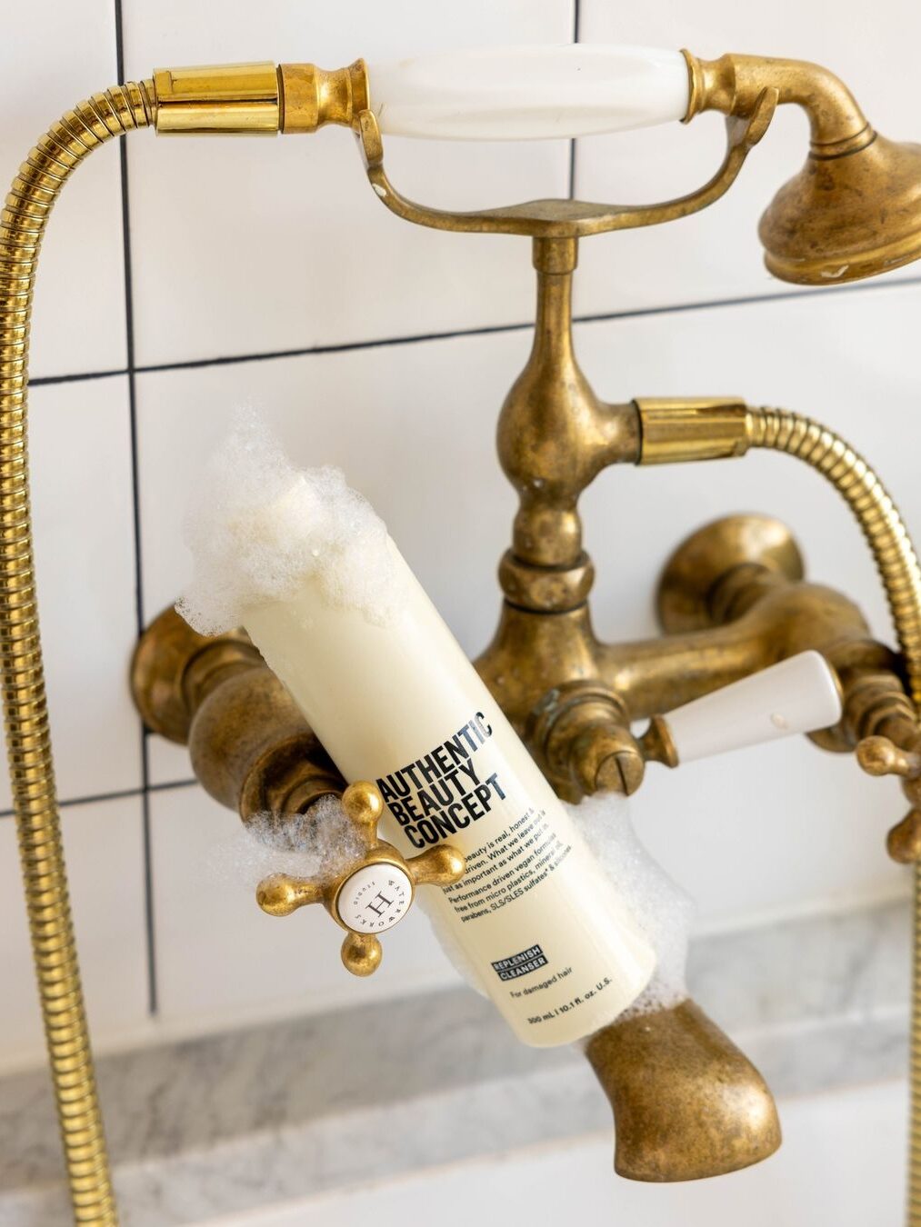 An Authentic Beauty Concept shampoo wedged in between a bathtub's taps. 