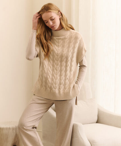 A person wearing a beige cable-knit sweater and matching pants stands beside an off-white chair in a softly lit room.