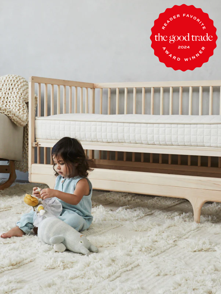 A child sits on a rug playing with toy animals in front of a wooden crib. A "Reader Favorite The Good Trade 2024 Award Winner" badge is visible in the top right corner.