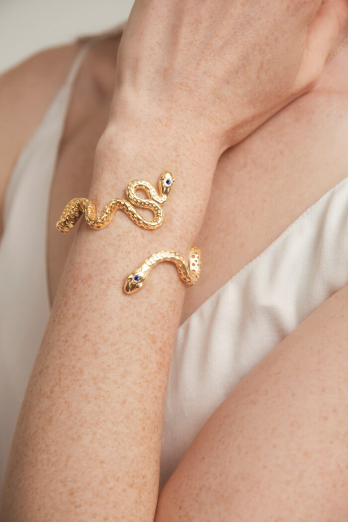 Close-up of a snake-shaped gold bracelet wrapped around a person's wrist, worn with a white sleeveless top.