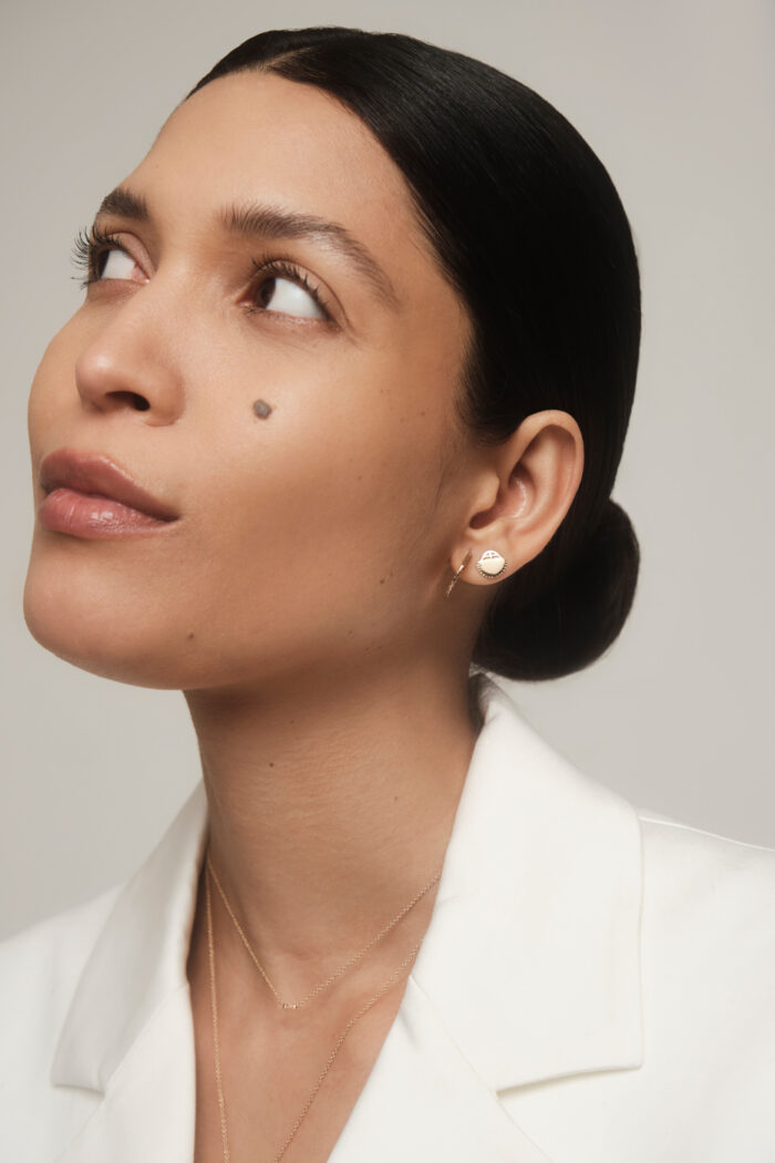 Person with slicked-back hair, wearing a white jacket, looking upwards. They have small earrings, a mole on their cheek, and a delicate necklace.