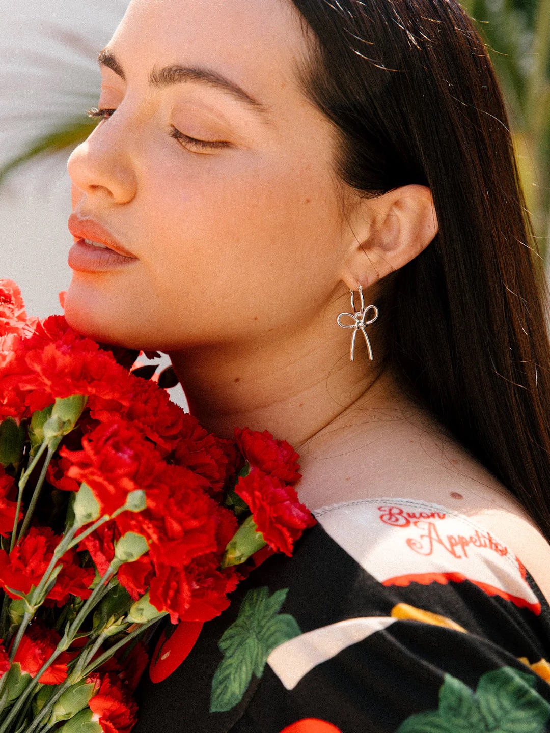 A woman with long dark hair holds a bouquet of red carnations, eyes closed, wearing a dress with "Bon Appétit" embroidered on the shoulder.