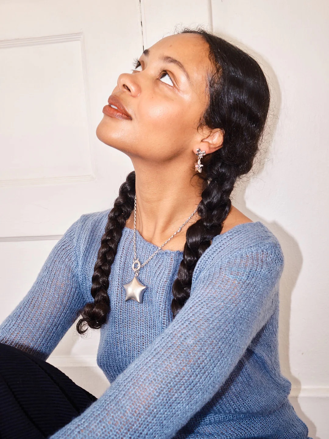 Person with braided hair gazes upwards, wearing a blue sweater and star-shaped jewelry.