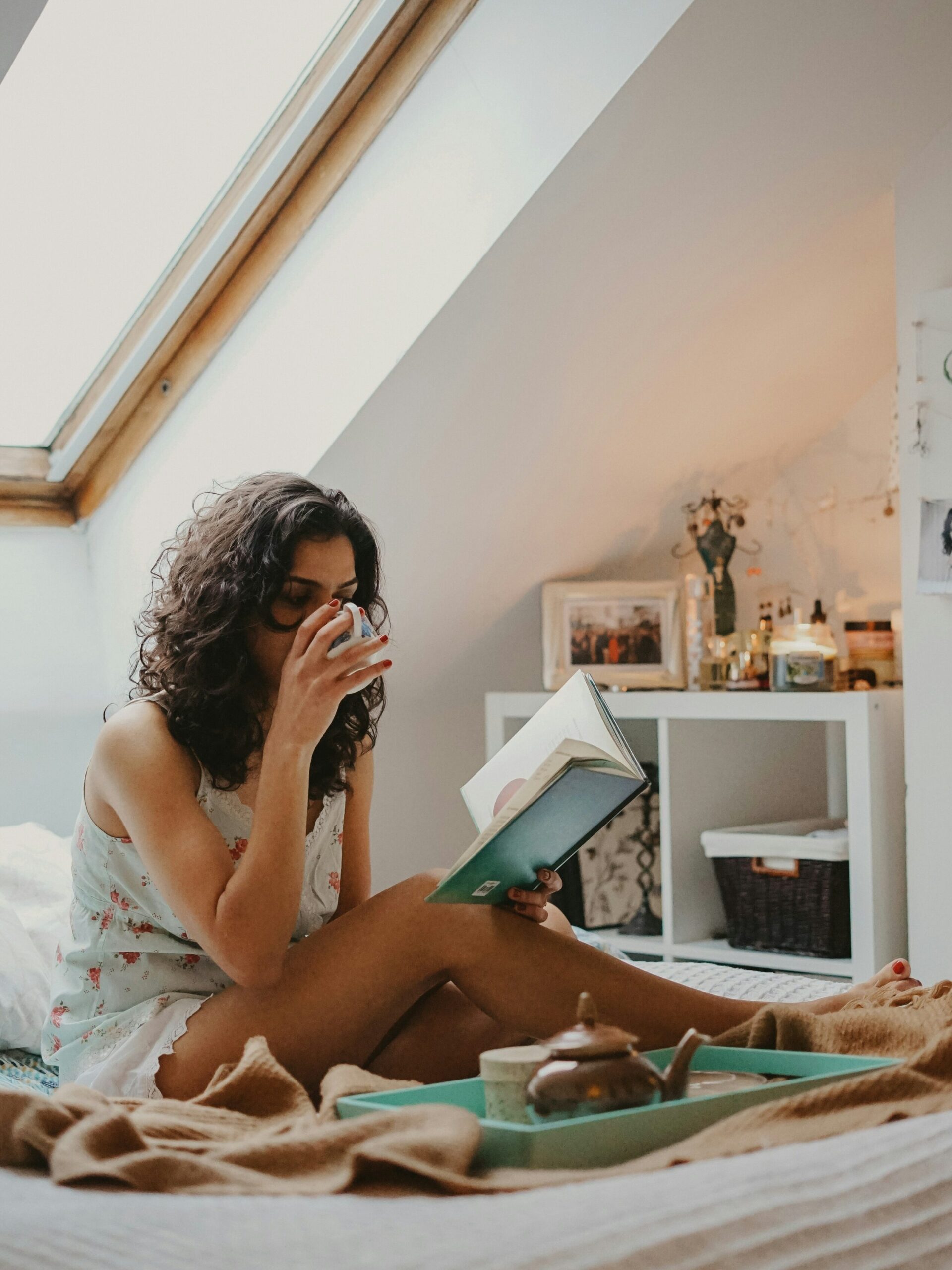 A model seated on a bed, drinking tea and reading their journal. 