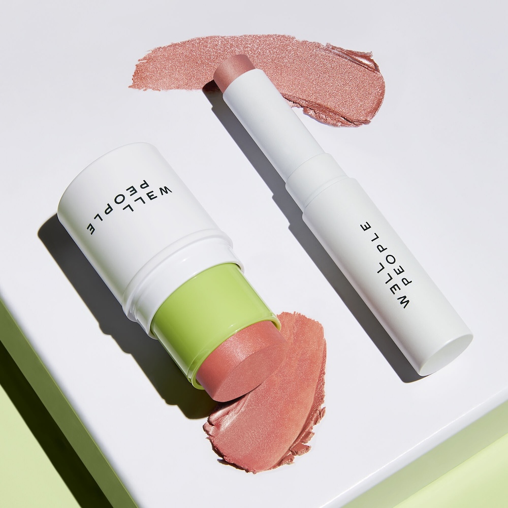 Two Well People cosmetic sticks on a white surface, with light pink swatches beside each; one in a white container with green cap, the other slim and white.